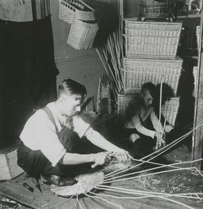876634 Afbeelding van een man en een jongen die rieten voorwerpen (o.a. manden en kinderzitjes) aan het vlechten zijn, ...